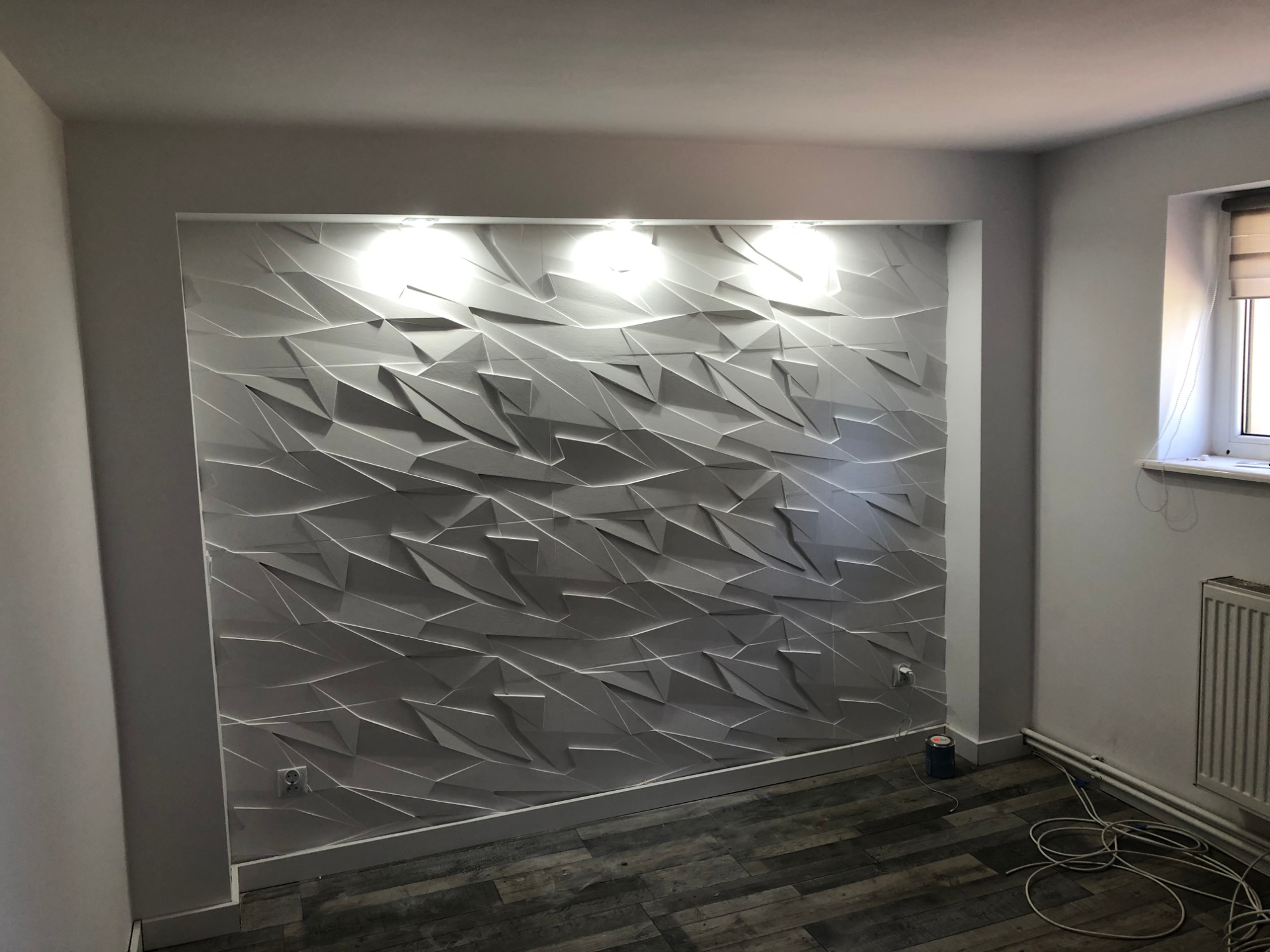 Pokój po remoncie: ściana z dekoracyjnymi panelami 3D o geometrycznym wzorze, oświetlona od góry, z widocznymi przewodami na podłodze.