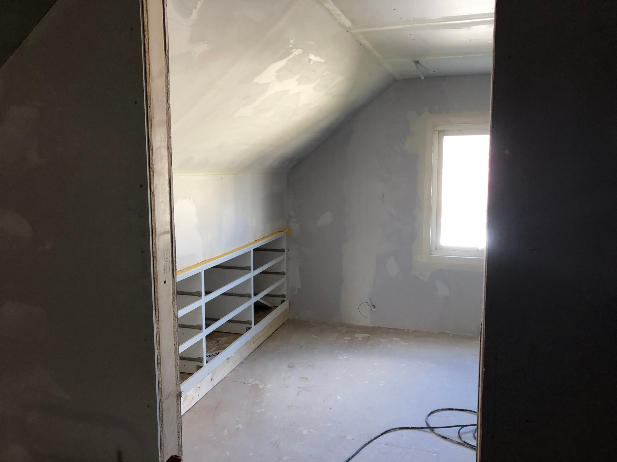 Poddasze w trakcie remontu: ściany po gładziowaniu, widoczne niedoskonałości, wbudowany regał z białymi półkami, kabel elektryczny na podłodze.