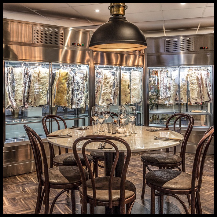 Restauracja z marmurowym stołem i krzesłami, w tle chłodnie z dojrzewającym mięsem, oświetlone ciepłym światłem lampy.
