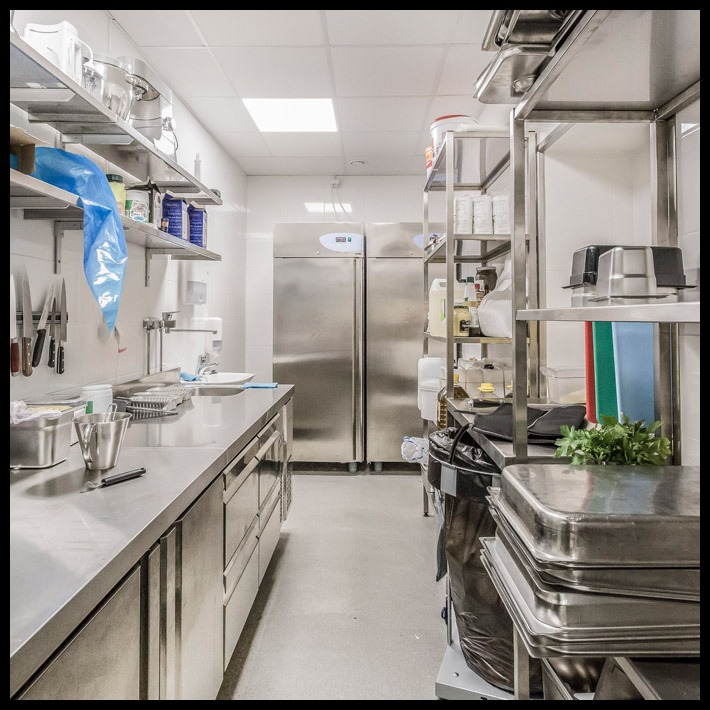 Wnętrze profesjonalnej kuchni z nierdzewnymi blatami, szafkami, półkami i dwoma dużymi lodówkami, widoczne akcesoria kuchenne i zioła na półce.