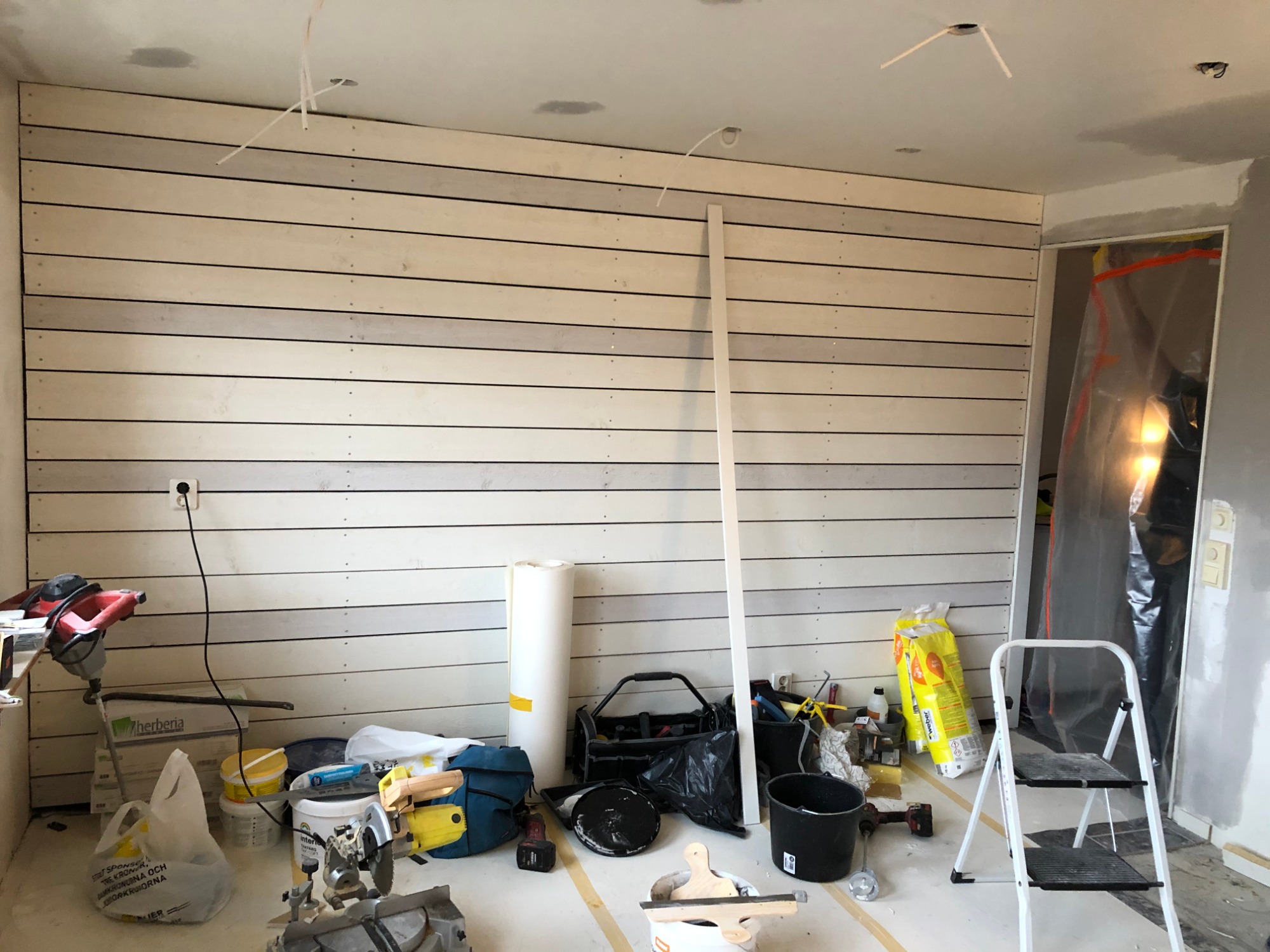 Ściana wykończona panelami boazeryjnymi w trakcie malowania, widoczne narzędzia i materiały budowlane, zabezpieczone folią drzwi.