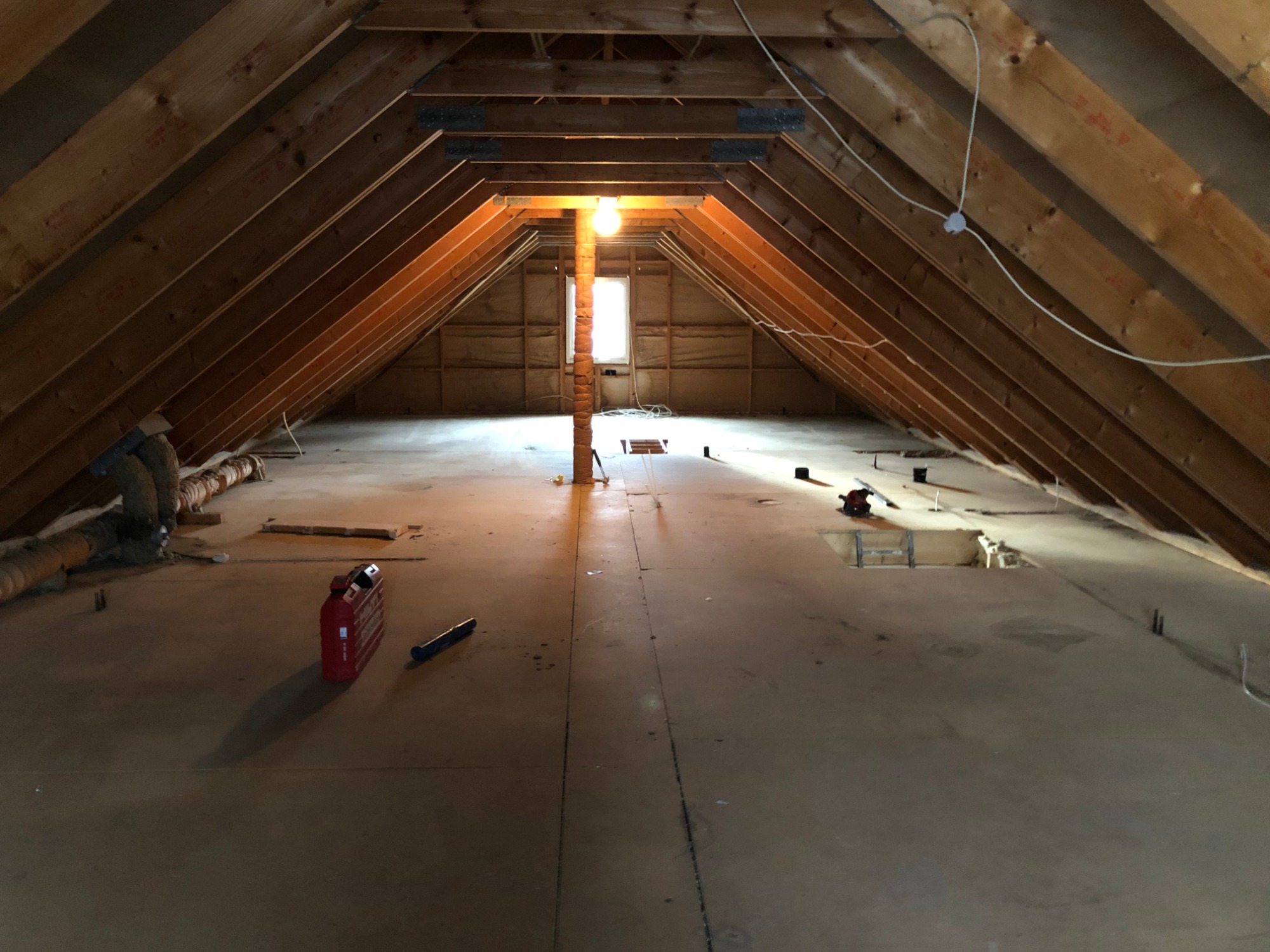 Poddasze w trakcie adaptacji: widoczne drewniane elementy konstrukcyjne dachu, izolacja rur, czerwona skrzynka narzędziowa i otwory w podłodze przygotowane pod instalacje.