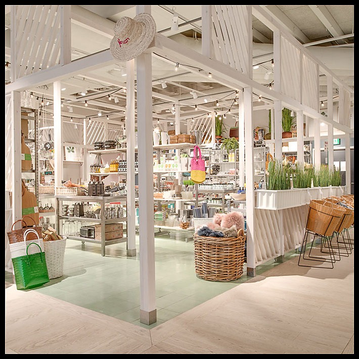 Jasne wnętrze sklepu z artykułami wyposażenia domu, biała drewniana konstrukcja, półki z różnorodnymi produktami, wiklinowe kosze i słomkowy kapelusz jako element dekoracyjny.