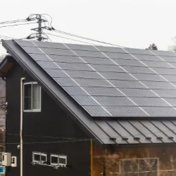 Dom jednorodzinny z ciemną elewacją i panelami fotowoltaicznymi na dachu, widok z boku, w tle linia energetyczna i zabudowa.