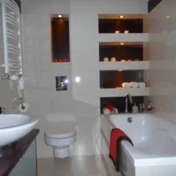 Nowoczesna łazienka z białą armaturą: umywalka nablatowa, toaleta, wanna z czerwoną szmatką, grzejnik drabinkowy, wnęki z oświetleniem i dekoracjami.