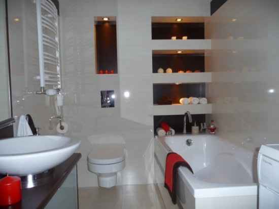 Nowoczesna łazienka z białą armaturą: umywalka nablatowa, toaleta, wanna z czerwoną szmatką, grzejnik drabinkowy, wnęki z oświetleniem i dekoracjami.