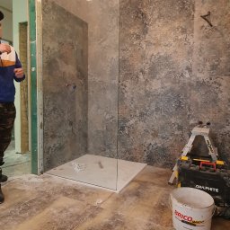 Piotr Bud - Montaż kabiny prysznicowej typu walk-in z szarą, wzorzystą okładziną ścienną. Widoczne narzędzia, drabina i wiadro z klejem. Podłoga zabezpieczona przed zabrudzeniem.