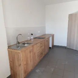 Nowoczesna, jasna kuchnia z drewnianymi szafkami, metalowym zlewem, czarnymi lampami wiszącymi i szarymi płytkami podłogowymi z widocznymi śladami butów.