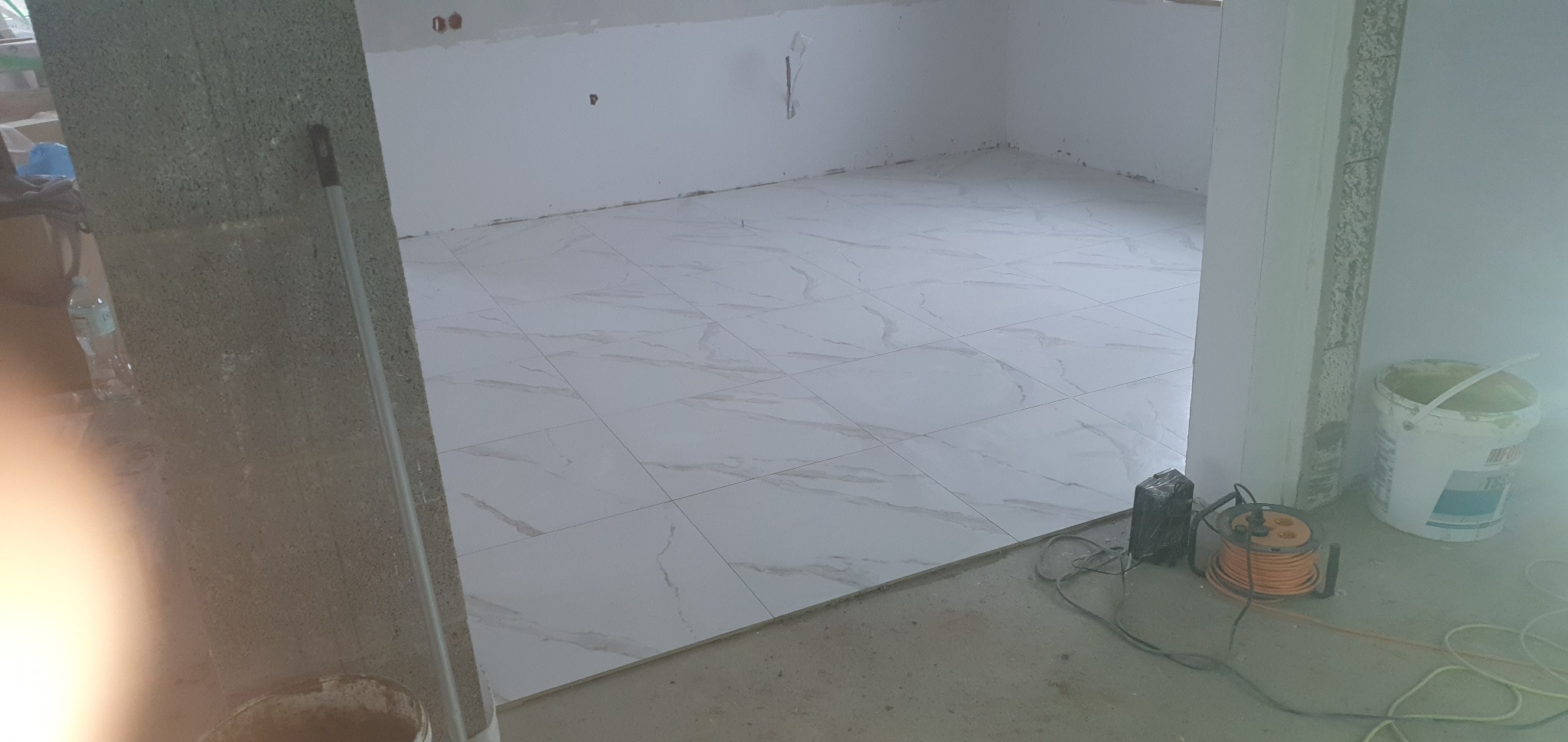 Ułożona podłoga z białych płytek imitujących marmur w pustym pomieszczeniu w trakcie remontu, widoczny kabel przedłużający i wiadro z farbą.
