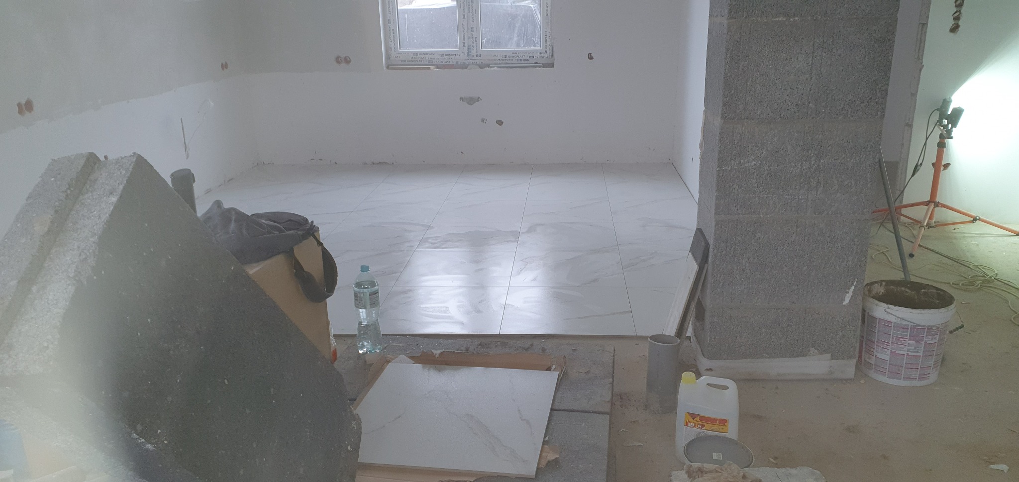 Podłoga wyłożona jasnymi płytkami imitującymi marmur w trakcie prac wykończeniowych, widoczne narzędzia, wiadro z zaprawą, butelka wody i nieotwarte paczki płytek.