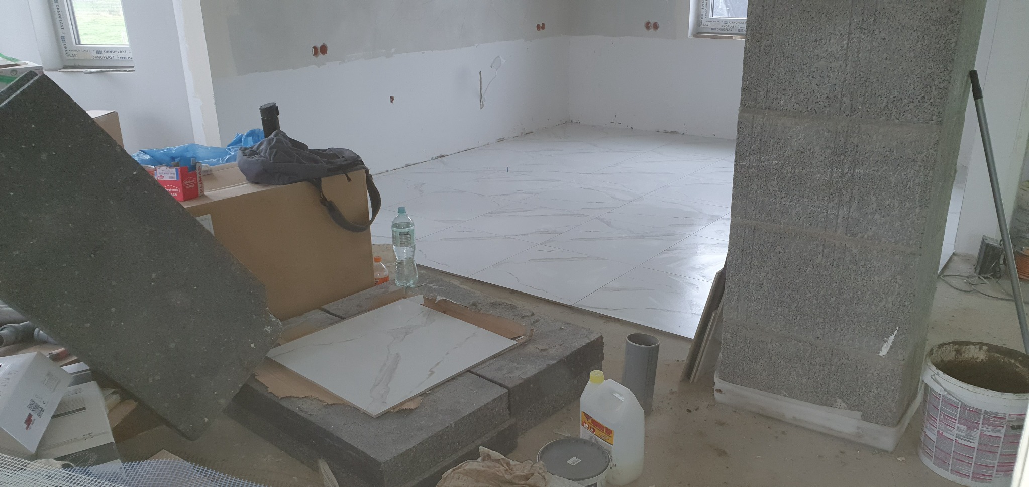 Układanie białych płytek podłogowych z szarym wzorem w trakcie remontu, widoczne materiały budowlane i narzędzia pracy.