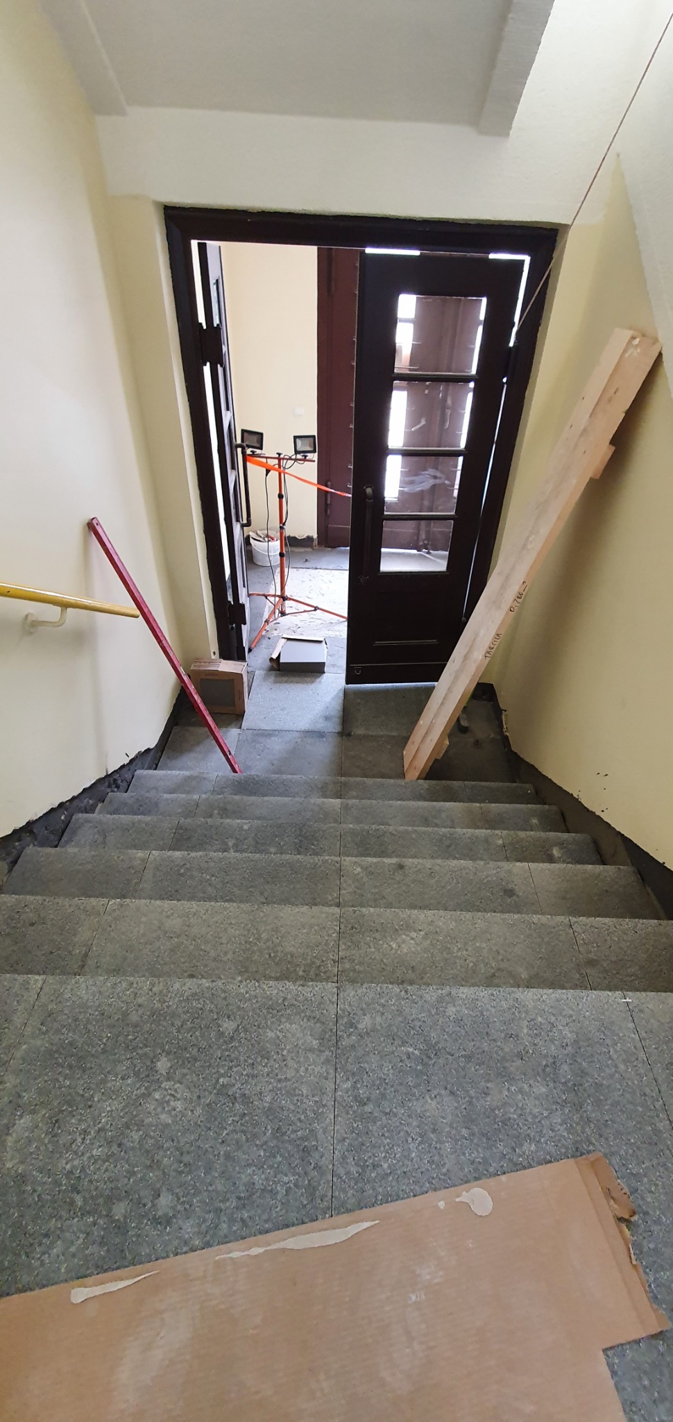 Schody wyłożone szarą, granitową płytką w trakcie prac wykończeniowych, widoczne narzędzia i oświetlenie budowlane, deski zabezpieczające ściany.