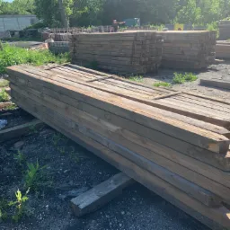 Stosy impregnowanych, kwadratowych bali drewnianych na placu składowym, z widoczną osobą w tle wskazującą kierunek.