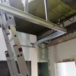 Montaż metalowej konstrukcji sufitu podwieszanego z widoczną drabina aluminiową i zieloną izolacją w tle, pomieszczenie w stanie surowym z widocznymi instalacjami i śladami szpachlowania.