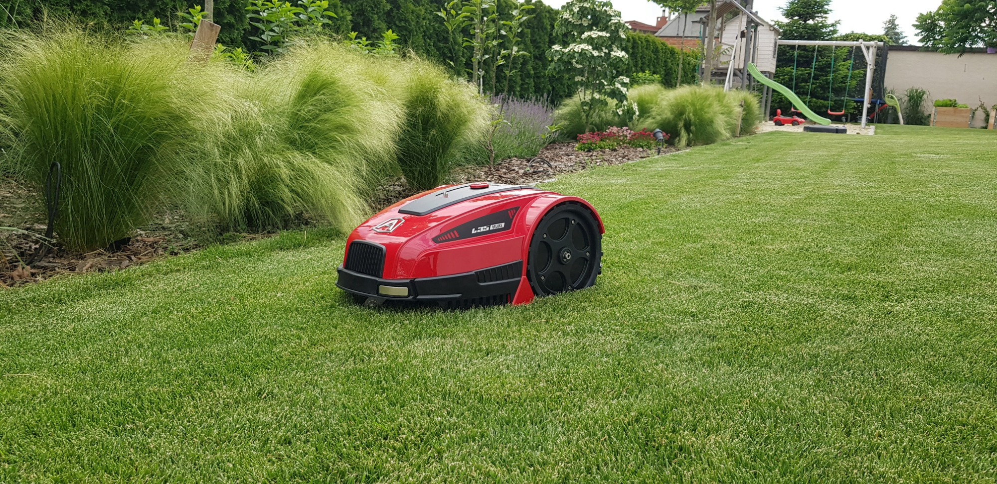 Czerwony robot koszący L55 Deluxe na idealnie przystrzyżonym trawniku, w tle ozdobne trawy, plac zabaw i zadbana zieleń.