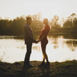 Para trzymająca się za ręce w Wilczkowicach na tle jeziora podczas zachodu słońca, kobieta w czerwonej sukience w ciąży, sylwetki na tle jasnego światła.