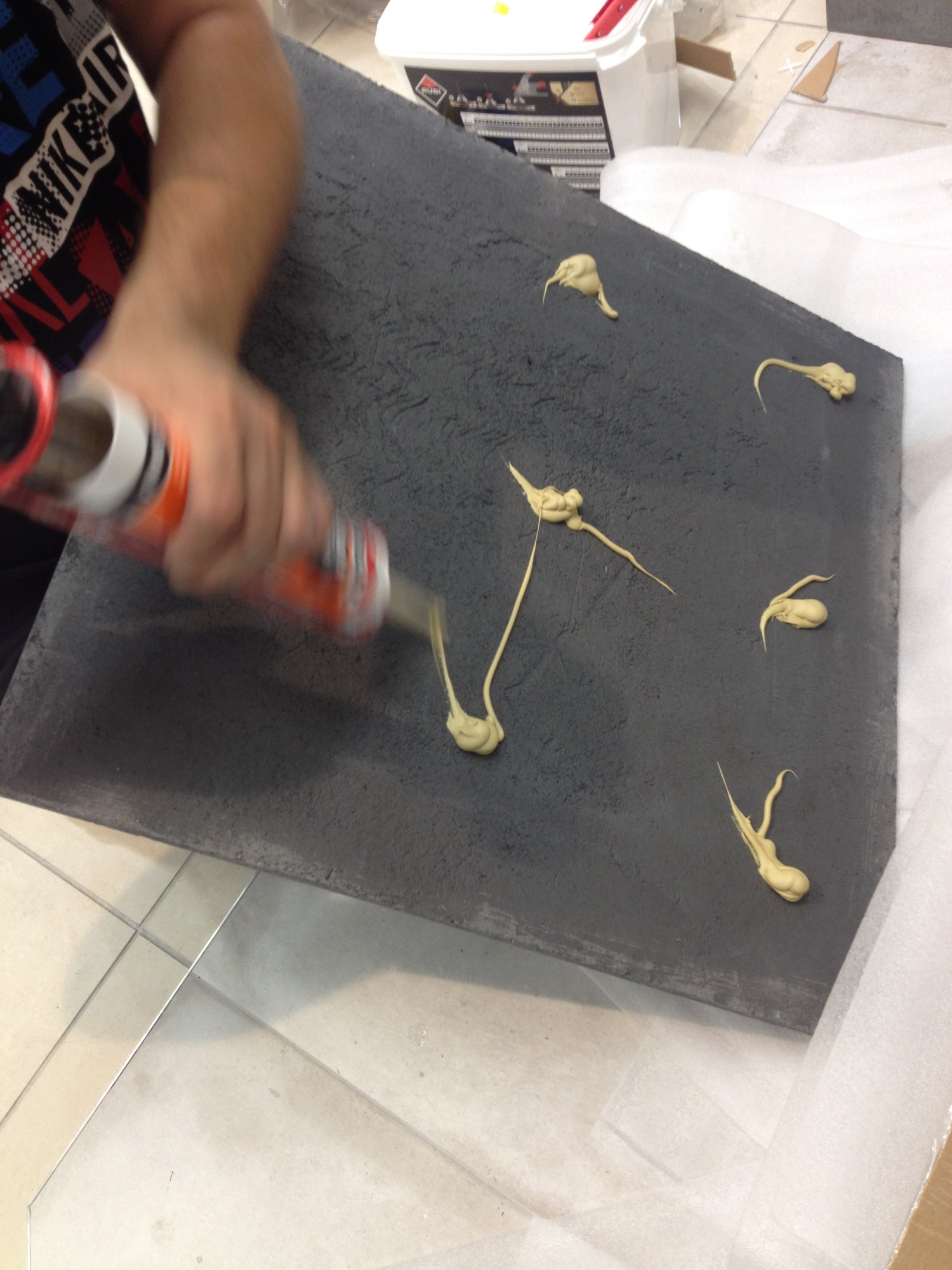 Nakładanie kleju montażowego na szarą płytę betonową z wyraźną fakturą, widoczna ręka z aplikatorem i klej w kilku miejscach na powierzchni płyty.