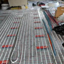 Montaż rur grzewczych podłogowych na macie z rastrem, widoczne narzędzia i inne elementy instalacji w surowym pomieszczeniu.