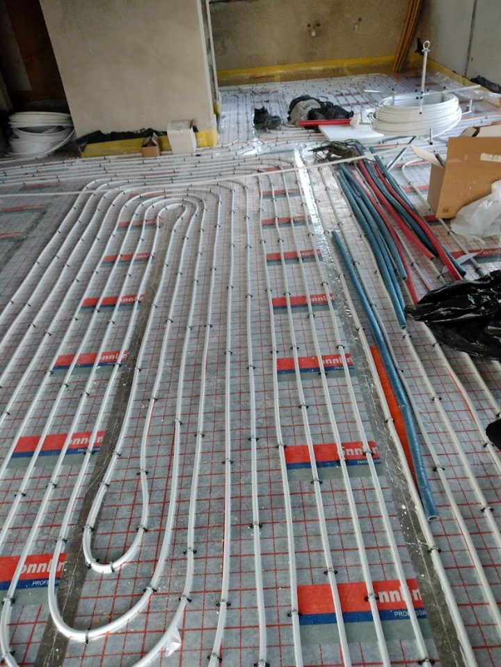 Montaż rur grzewczych podłogowych na macie z rastrem, widoczne narzędzia i inne elementy instalacji w surowym pomieszczeniu.