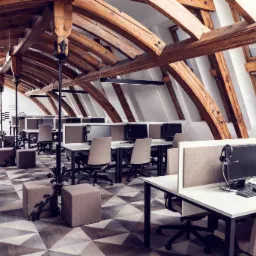 Przestronne biuro z drewnianą konstrukcją stropu, stanowiska pracy wyposażone w monitory i zestawy słuchawkowe, wzorzysta wykładzina podłogowa.