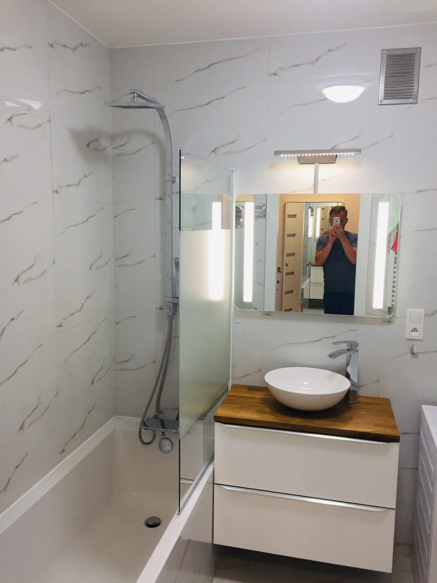 Nowoczesna łazienka z białymi marmurkowymi płytkami, kabiną prysznicową ze szklaną ścianką, umywalką nablatową na drewnianym blacie i lustrem z podświetleniem LED, widoczny refleks fotografa...