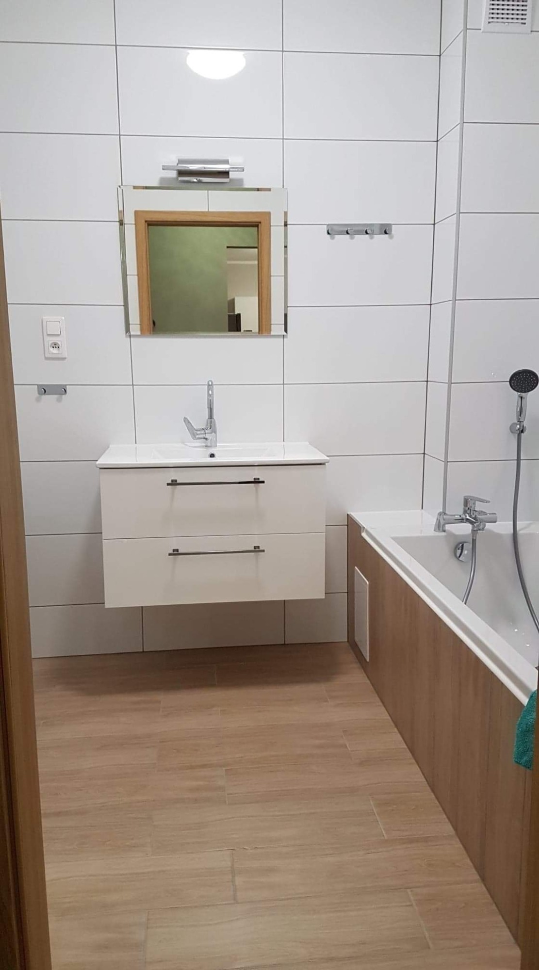 Nowoczesna łazienka z białą, wiszącą szafką pod umywalką, lustrem w drewnianej ramie i wanną obudowaną panelami imitującymi drewno, wykończona białymi płytkami i drewnopodobną podłogą.