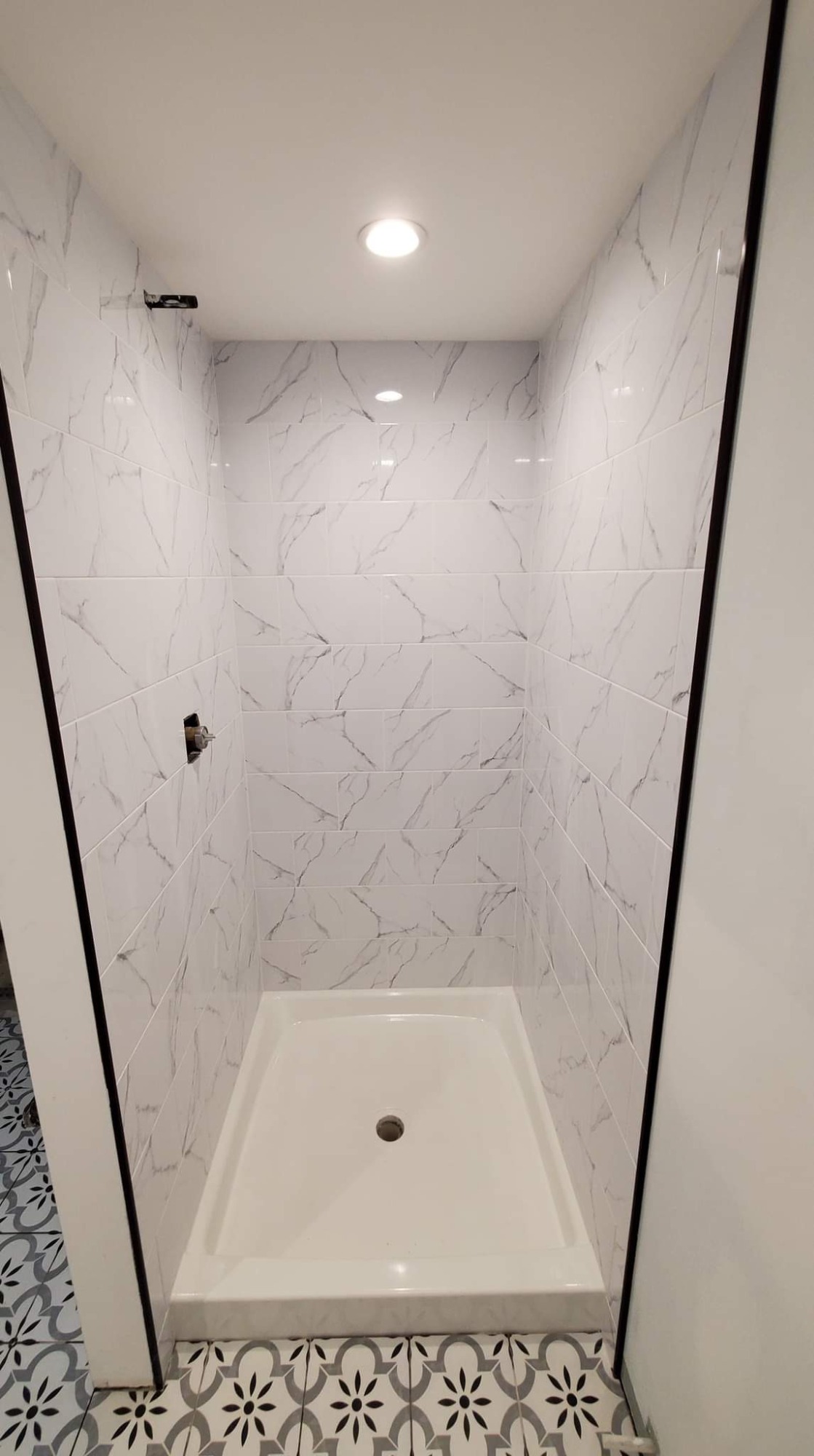 Nowoczesna kabina prysznicowa z białym brodzikiem i marmurowymi płytkami ściennymi, wykończona czarnymi listwami, z widocznymi elementami instalacji wodnej i dekoracyjną podłogą w kwiatowy wzór.