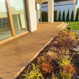 Beżowy taras z desek kompozytowych przy nowoczesnym domu z dużymi oknami, widok na ogród z tujami.