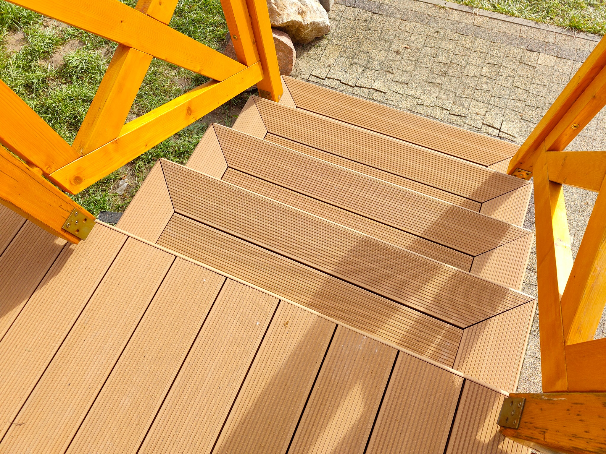 Schody z desek kompozytowych prowadzące na taras, z elementami drewnianej konstrukcji w kolorze pomarańczowym, widoczne kamienie i kostka brukowa.