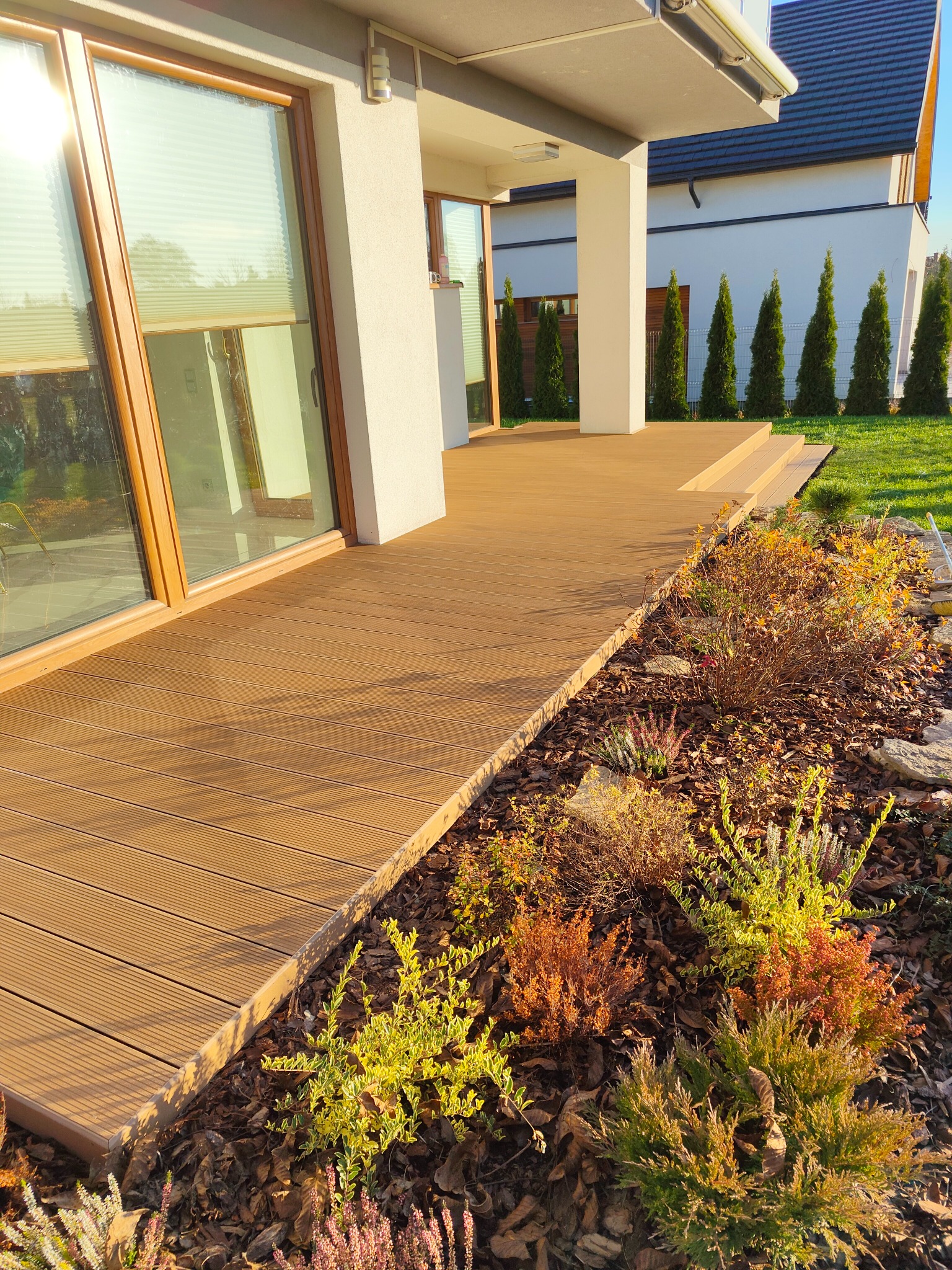 Beżowy taras z desek kompozytowych przy nowoczesnym domu z dużymi oknami, widok na ogród z tujami.