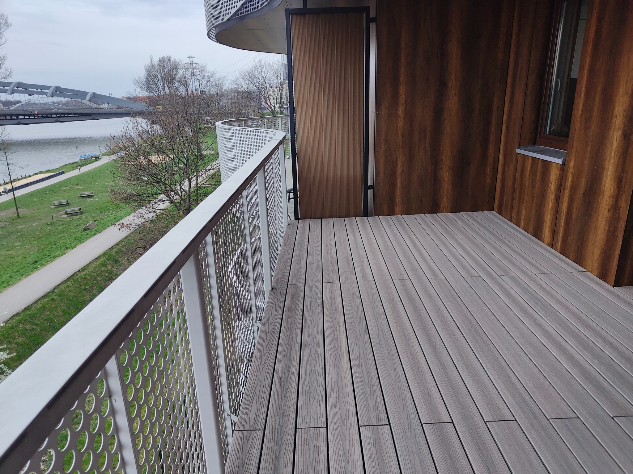 Szary taras z desek kompozytowych z widokiem na rzekę i most; ażurowa, biała balustrada i drewniana ściana z oknem.
