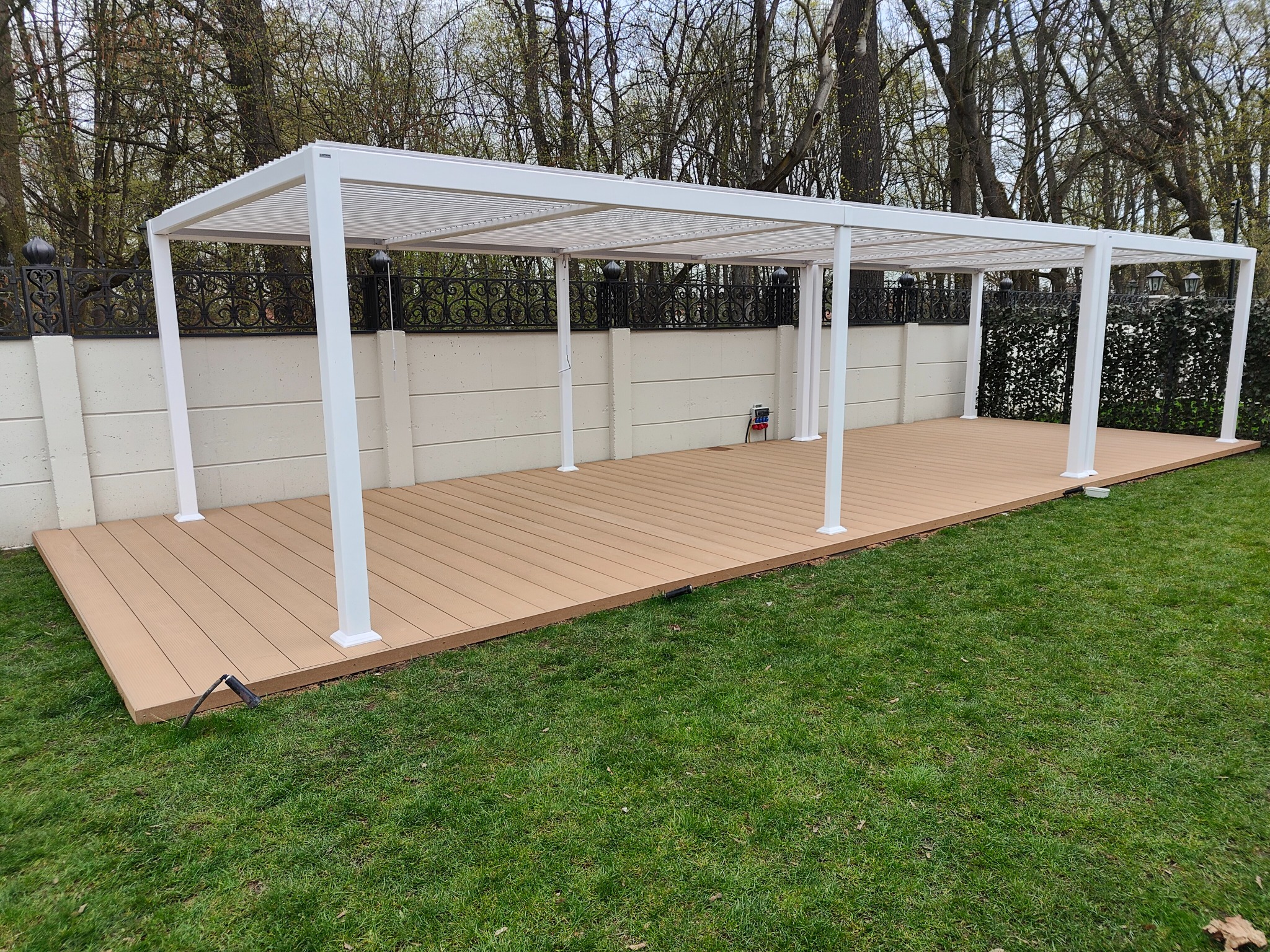 Biała, metalowa pergola z lamelowym dachem na tarasie z desek kompozytowych, widok na ogród z trawnikiem i ogrodzeniem.