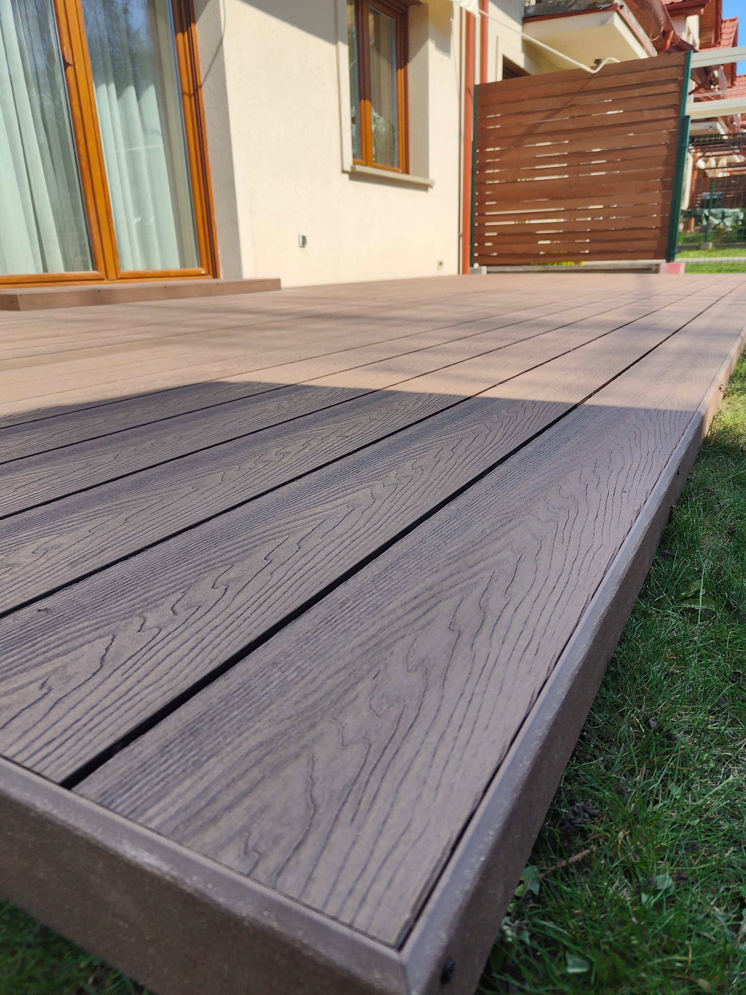 Nowoczesny taras kompozytowy w kolorze brązowym, z widoczną fakturą drewna, otoczony zielonym trawnikiem, przylegający do jasnego budynku z oknami w drewnianych ramach.