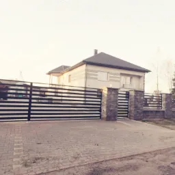 Nowoczesna, pozioma brama wjazdowa i ogrodzenie z czarnych, płaskich profili, osadzone na kamiennych słupkach, przed dwukondygnacyjnym domem z szarym dachem.