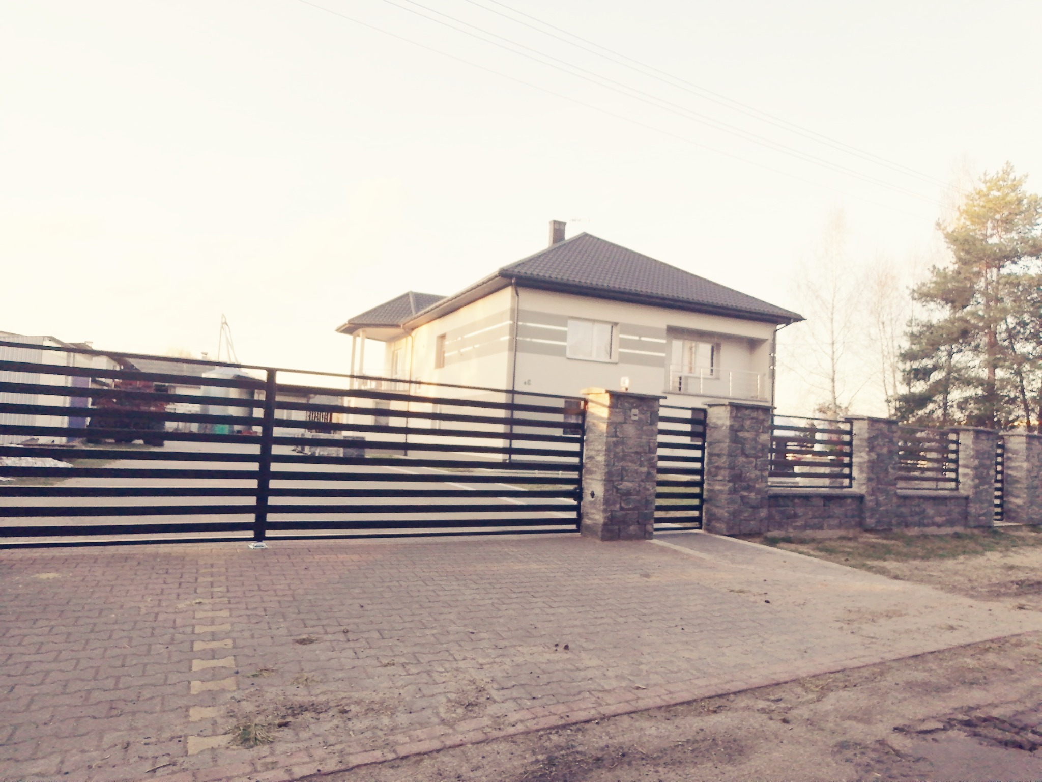 Nowoczesna, pozioma brama wjazdowa i ogrodzenie z czarnych, płaskich profili, osadzone na kamiennych słupkach, przed dwukondygnacyjnym domem z szarym dachem.