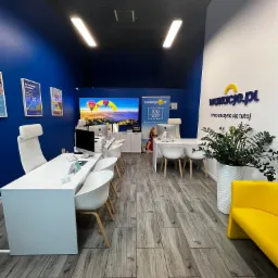 Wnętrze biura podróży z białymi meblami, niebieską ścianą, monitorami z motywem balonów i logo 'wakacje.pl'