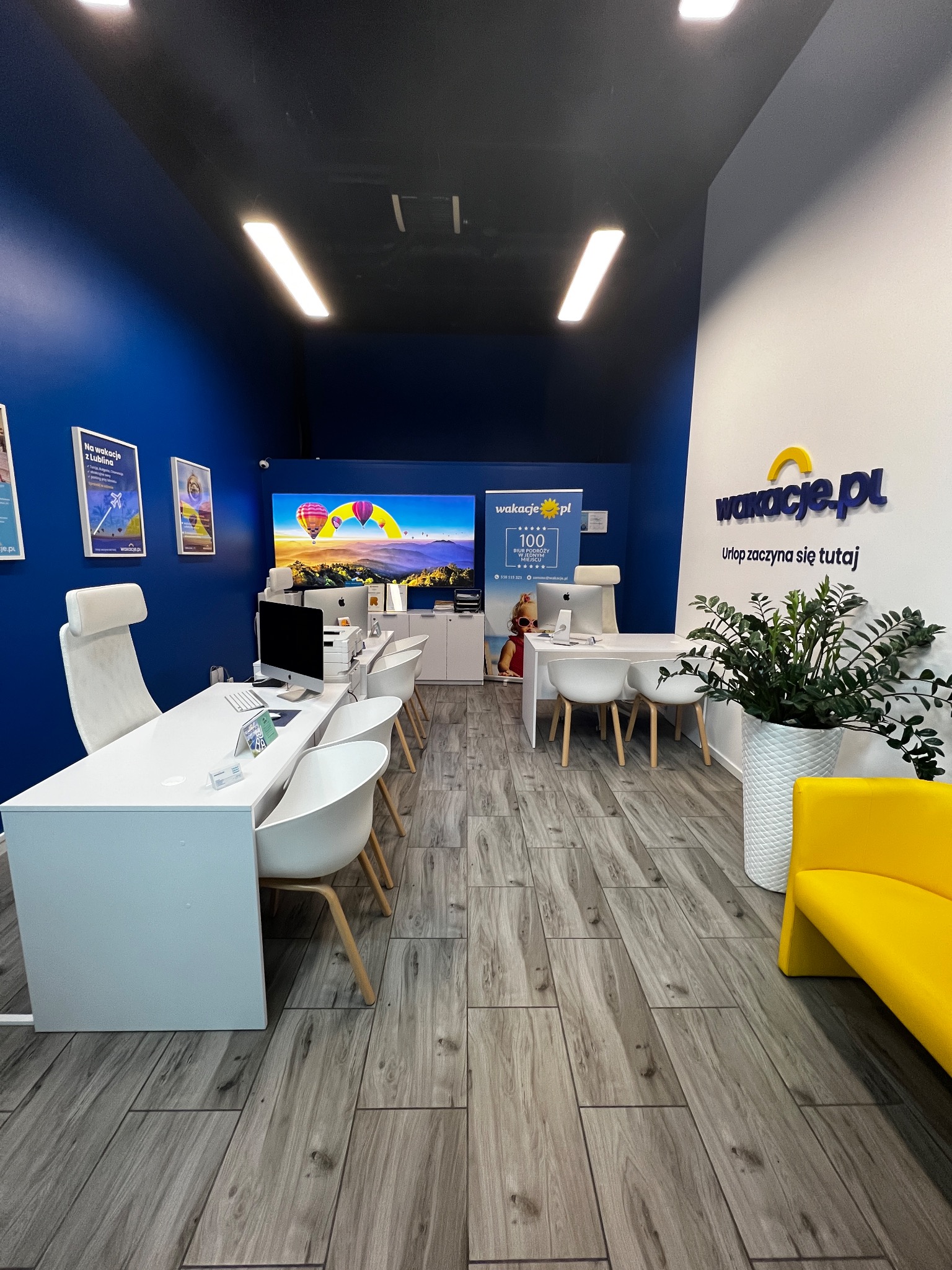 Wnętrze biura podróży z białymi meblami, niebieską ścianą, monitorami z motywem balonów i logo 'wakacje.pl'