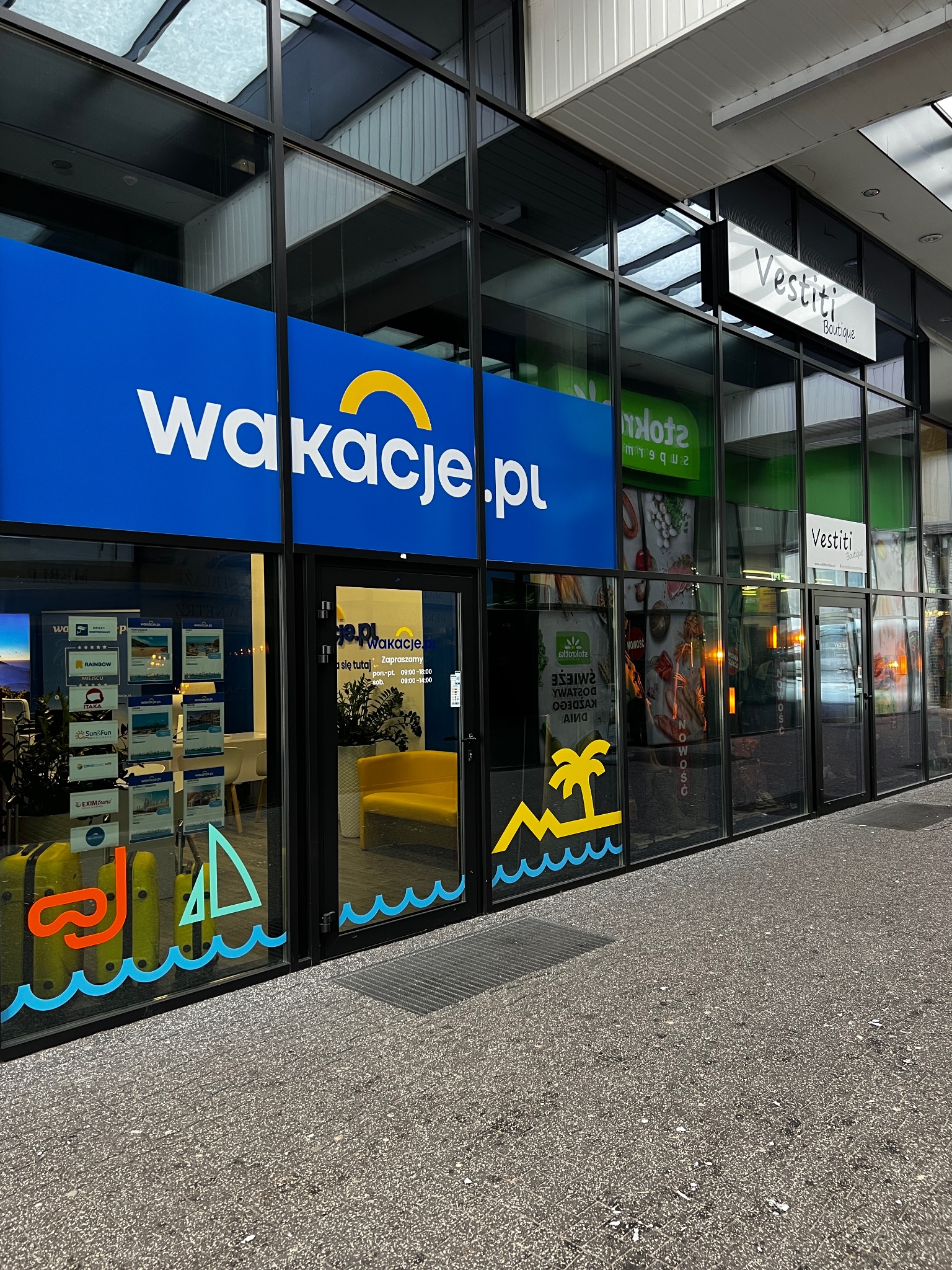 Fasada biura podróży Wakacje.pl z dużym niebieskim szyldem, grafikami wakacyjnymi na oknach i widocznym wnętrzem z żółtą sofą.