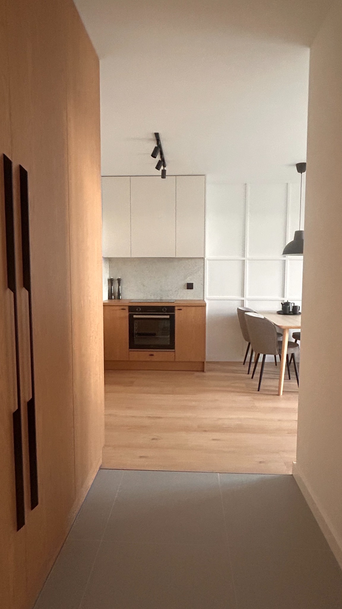 Nowoczesne wnętrze: jasna kuchnia z drewnianymi szafkami i szarymi płytkami podłogowymi, widok z korytarza. Minimalistyczny design, oświetlenie punktowe.