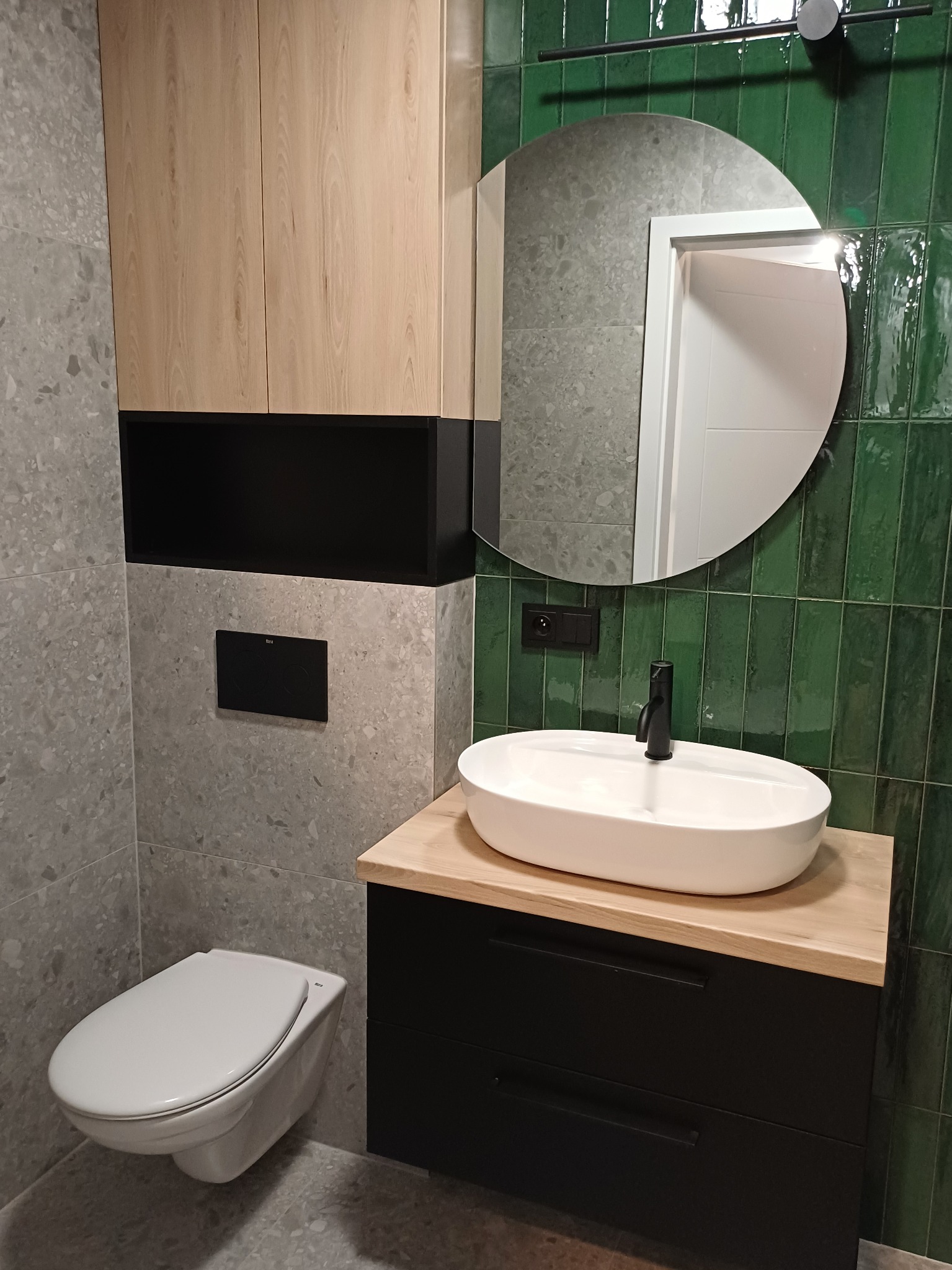 Nowoczesna łazienka w Gdańsku: szare płytki, zielona ściana z lustrem, podwieszana toaleta i szafka z umywalką. Minimalistyczny design, eleganckie wykończenie.