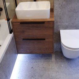 Bud-dud - Nowoczesna łazienka z wiszącą szafką pod umywalkę z drewnianymi szufladami i podświetleniem LED, biała umywalka nablatowa z czarną armaturą, toaleta podwieszana na tle szarej ściany...