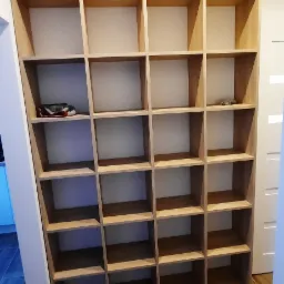 Wysoka, drewniana biblioteczka z pustymi półkami, w dolnej części na jednej z półek leżą okulary, a na innej czerwone buty sportowe.