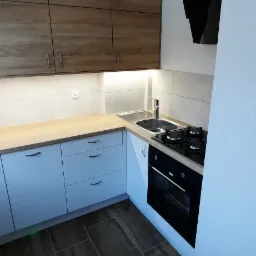 Nowoczesna kuchnia z białymi szafkami dolnymi i drewnopodobnymi górnymi, drewniany blat, płyta gazowa i czarny okap, brązowe płytki podłogowe imitujące drewno.