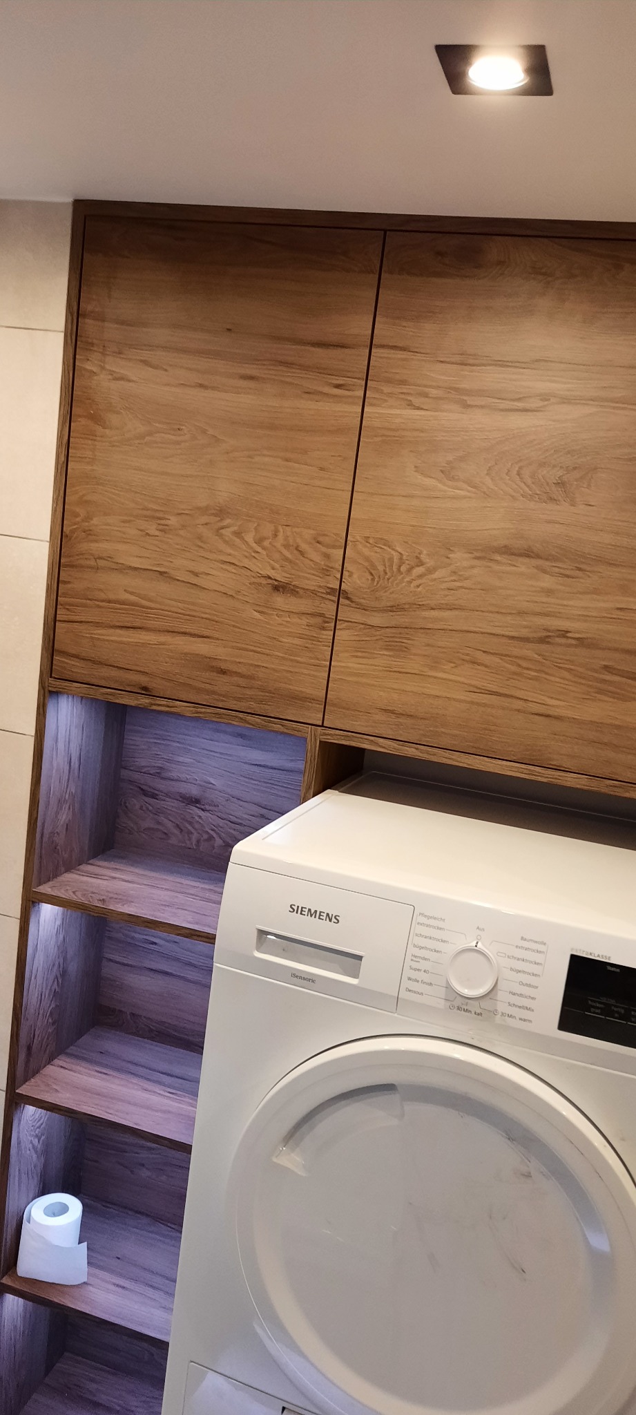 Nowoczesna łazienka z zabudową meblową z drewnopodobnymi frontami, wnęką z podświetlanymi półkami i pralką marki Siemens.