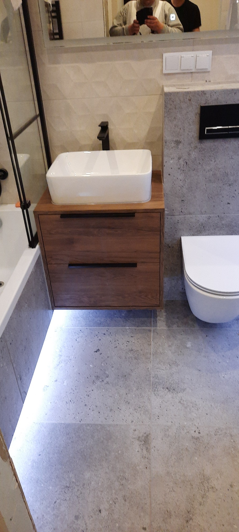 Nowoczesna łazienka z wiszącą szafką pod umywalkę z drewnianymi szufladami i podświetleniem LED, biała umywalka nablatowa z czarną armaturą, toaleta podwieszana na tle szarej ściany...