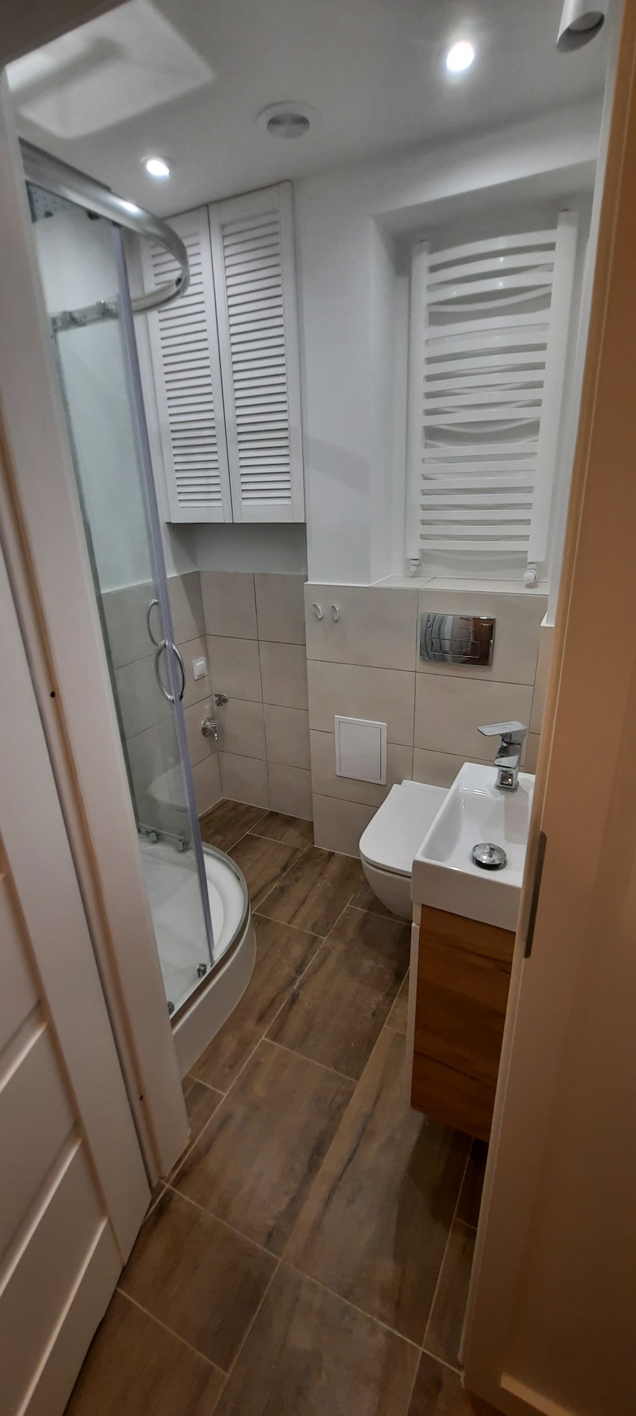 Nowoczesna łazienka po remoncie z kabiną prysznicową, toaletą, umywalką z szafką z drewnopodobnym frontem, grzejnikiem drabinkowym i szafką z żaluzjowymi drzwiczkami.