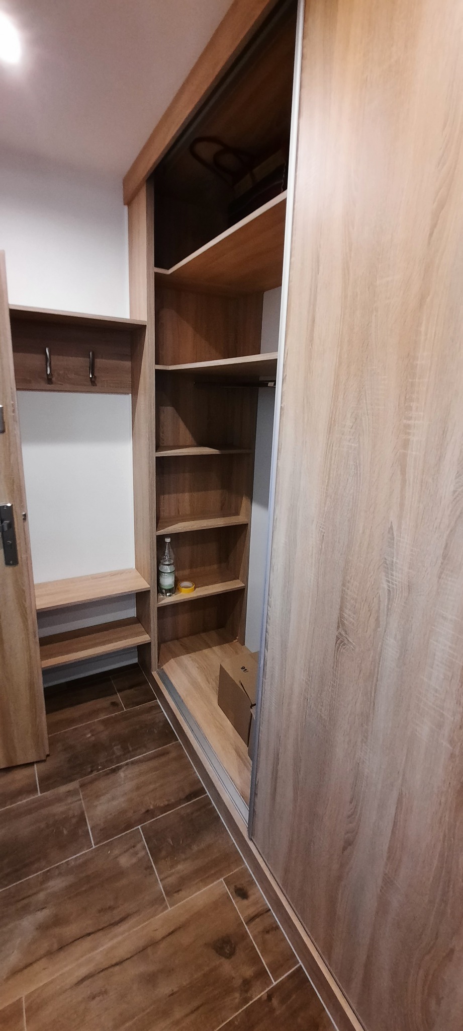 Wnętrze szafy przesuwnej z jasnego drewna z półkami, w rogu butelka wody mineralnej i taśma klejąca, obok wieszak z haczykami i siedziskiem, brązowe płytki na podłodze.