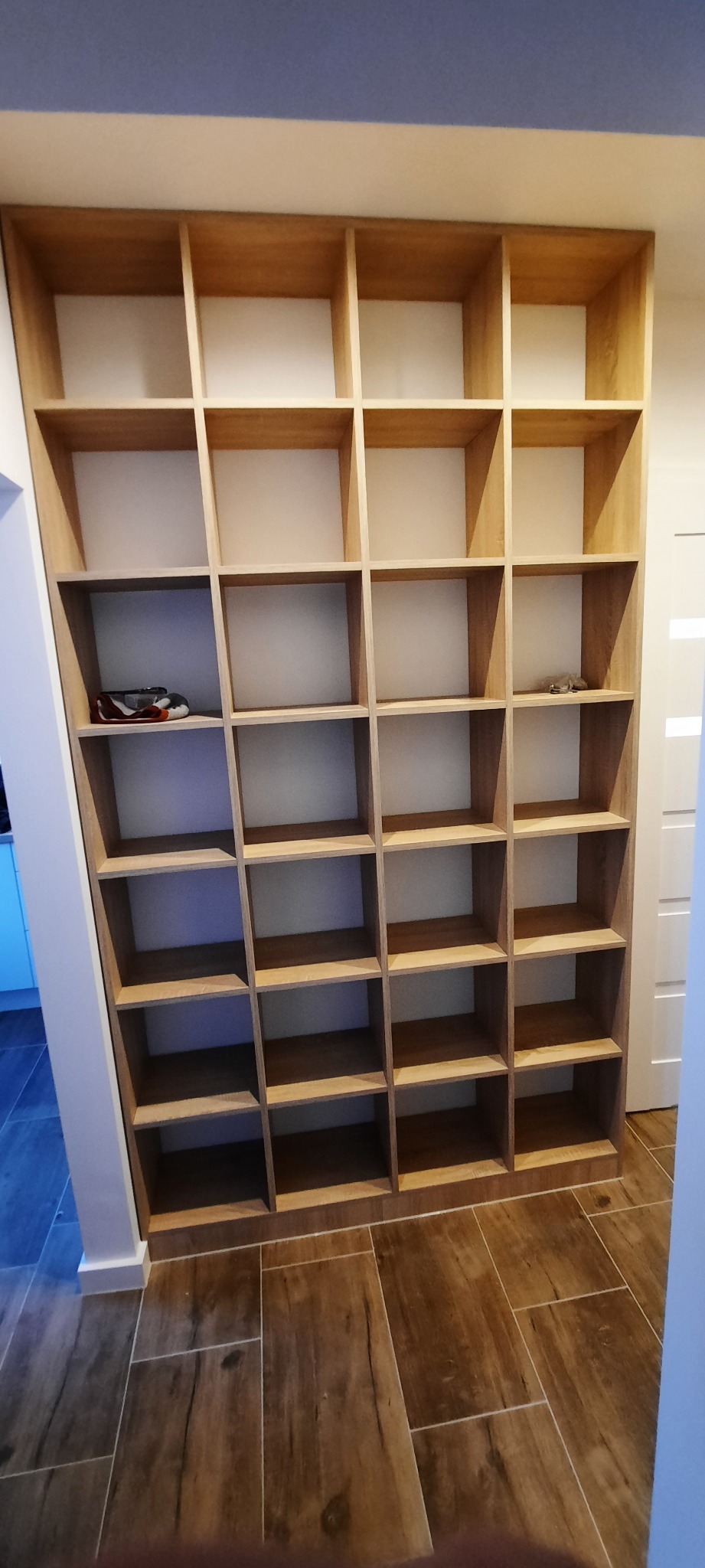 Wysoka, drewniana biblioteczka z pustymi półkami, w dolnej części na jednej z półek leżą okulary, a na innej czerwone buty sportowe.