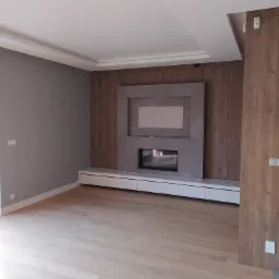 Salon z drewnianą podłogą, szarą ścianą i zabudową kominkową z szarą ramą na telewizor, drewnianymi panelami i białymi szafkami pod spodem.