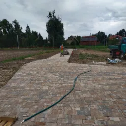 Świeżo ułożona, zakrzywiona ścieżka z kostki brukowej w odcieniach brązu i szarości, z zielonym wężem ogrodowym na jej powierzchni, prowadząca w kierunku pracownika w odblaskowej kamizelce...